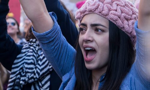 Woman wearing pink hat and blue shirt raising their arms at a protest.