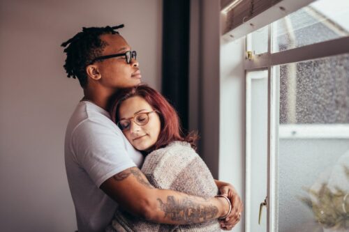Interracial couple hugging each other peacefully while standing near window. Black man embracing his white girlfriend at home.