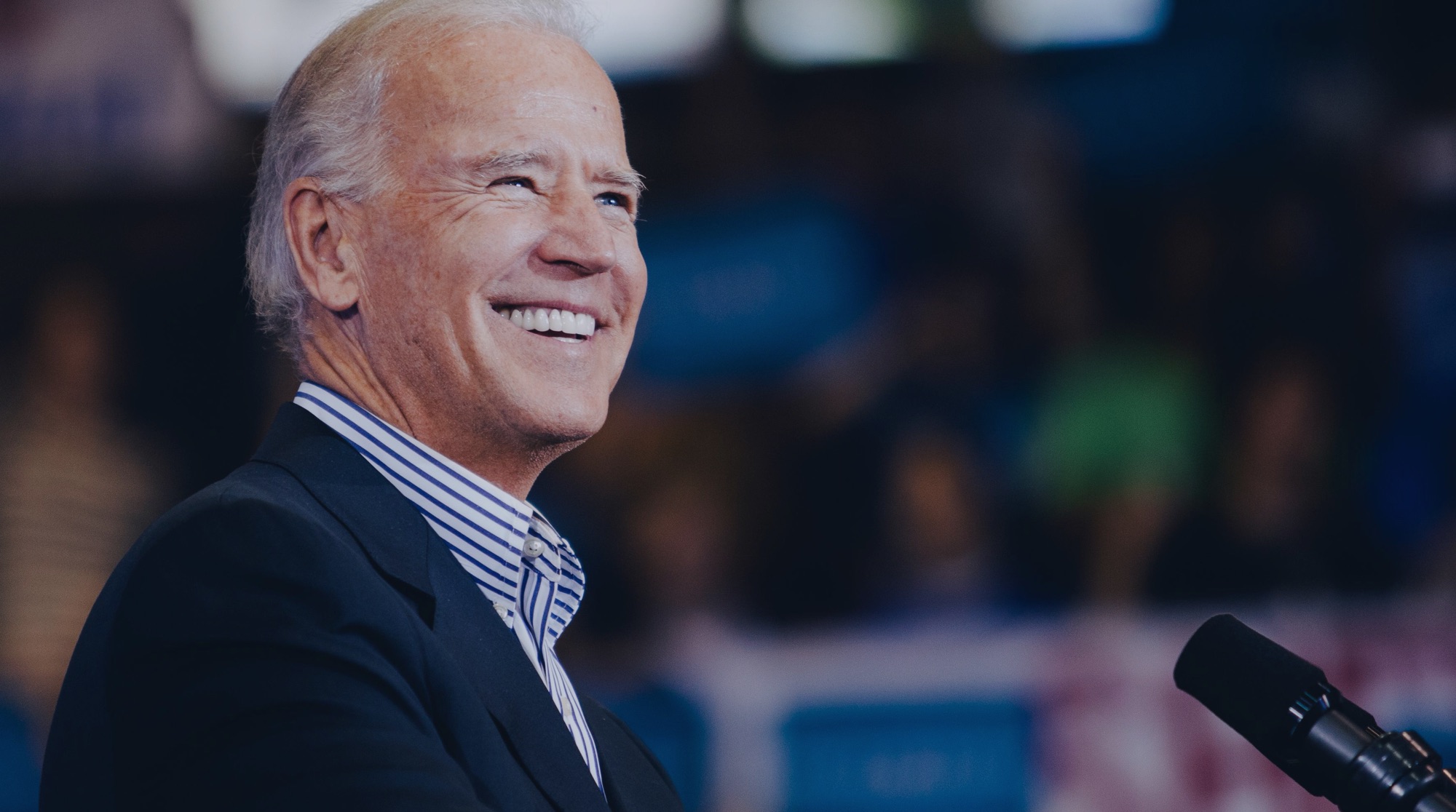Joe Biden smiling while standing at a podium, in front of a large crowd blurred in the background.