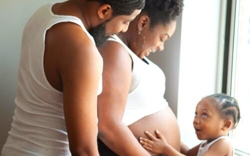 A Black father and mother stand up against each other, while their smiling child holds the mom's pregnant belly.