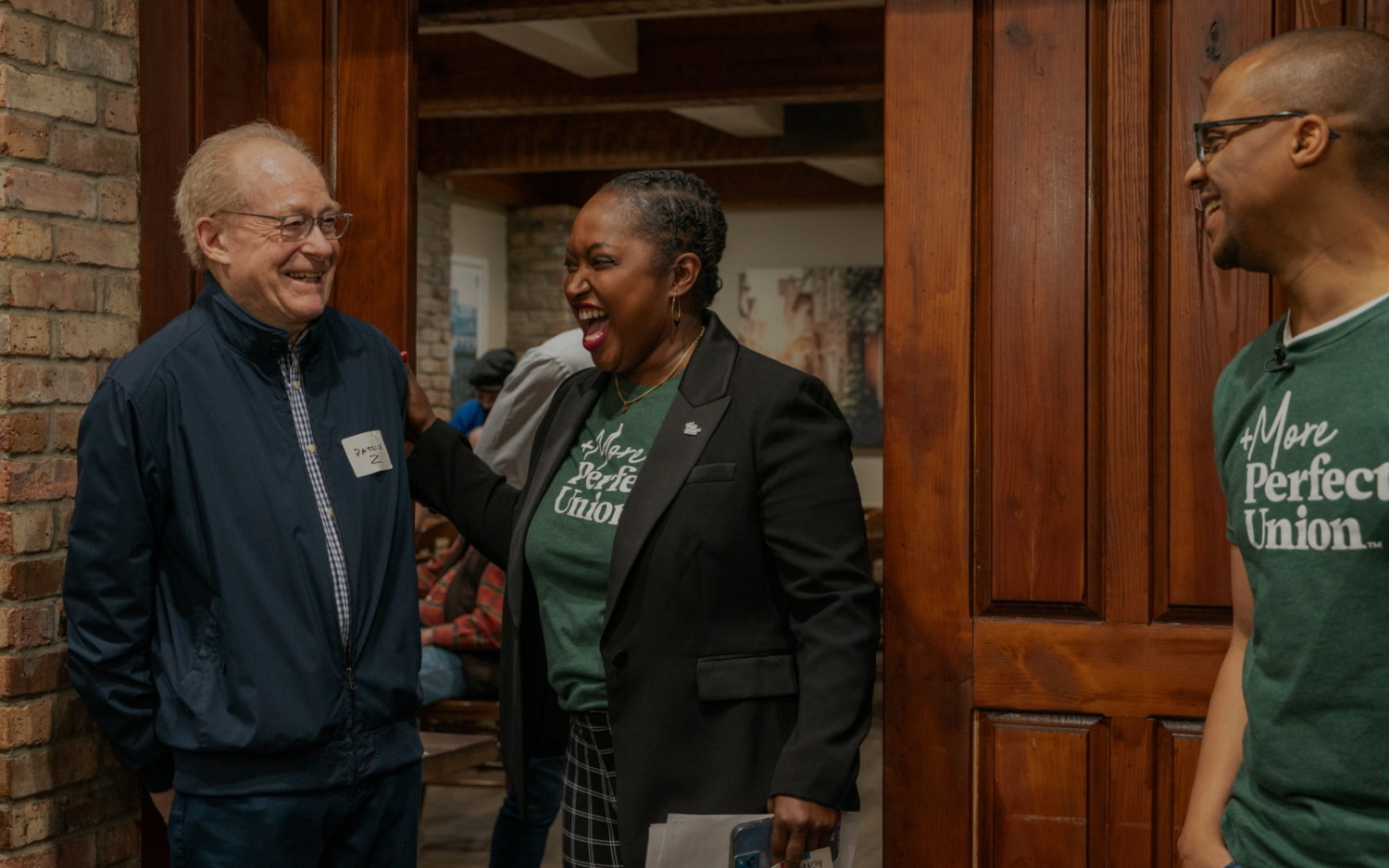 Black, woman Brickyard director and military veteran of +MPU, Mary Tobin places her hand while laughing joyfully on an older gentleman. They appear to be coming out of wood-stained doors with a bustling event happening behind them. Off to the side is Brickyard leader, Archie Nettles, who matches Mary in a green +MPU shirt and is seen smiling in their direction.