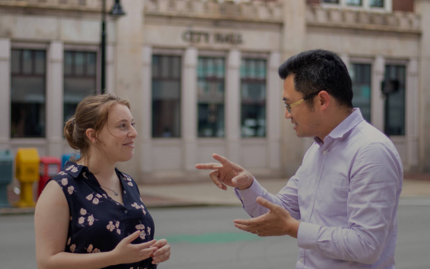 A man and woman talk animatedly in front of a city hall building.