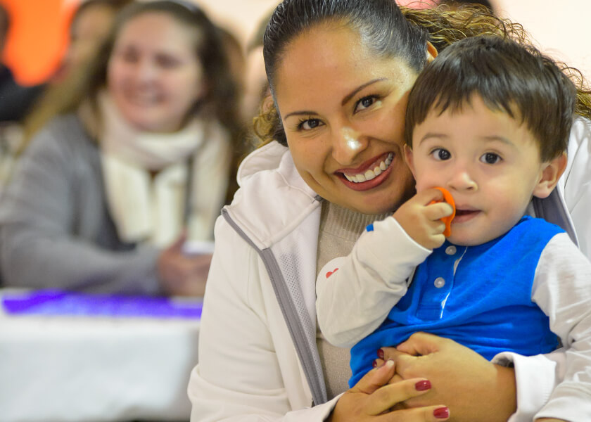 A toddler sits on its mother's lap while she smiles