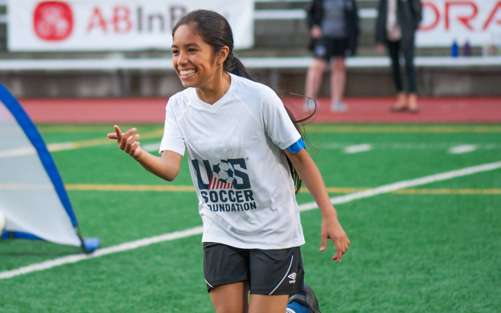 A middle-school-age, hispanic girl with olive skin and long, black hair in a pony-tail runs on a green soccer field. Her hair blows slightly in the wind and she has a big smile on her face. She points and is leaning forward as she plays soccer. Her shirt is white with the US Soccer Foundation logo printed on it. She has on navy shorts with a skinny, white stripe running alongside each thigh. One foot is lifted off the ground. She runs wearing black cleats and blue socks. A bright blue soccer net bleeds slightly into few on the right side of the image.