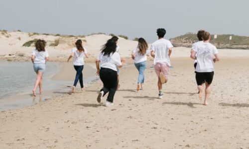 A group of six Diller Teen Fellows running away from the camera on a white sand beach during an overcast day.