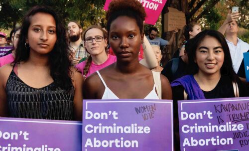 Latinx, Black and Asian-American women stand in protest holding reproductive freedom signs at a rally for Reproductive Freedom for All.