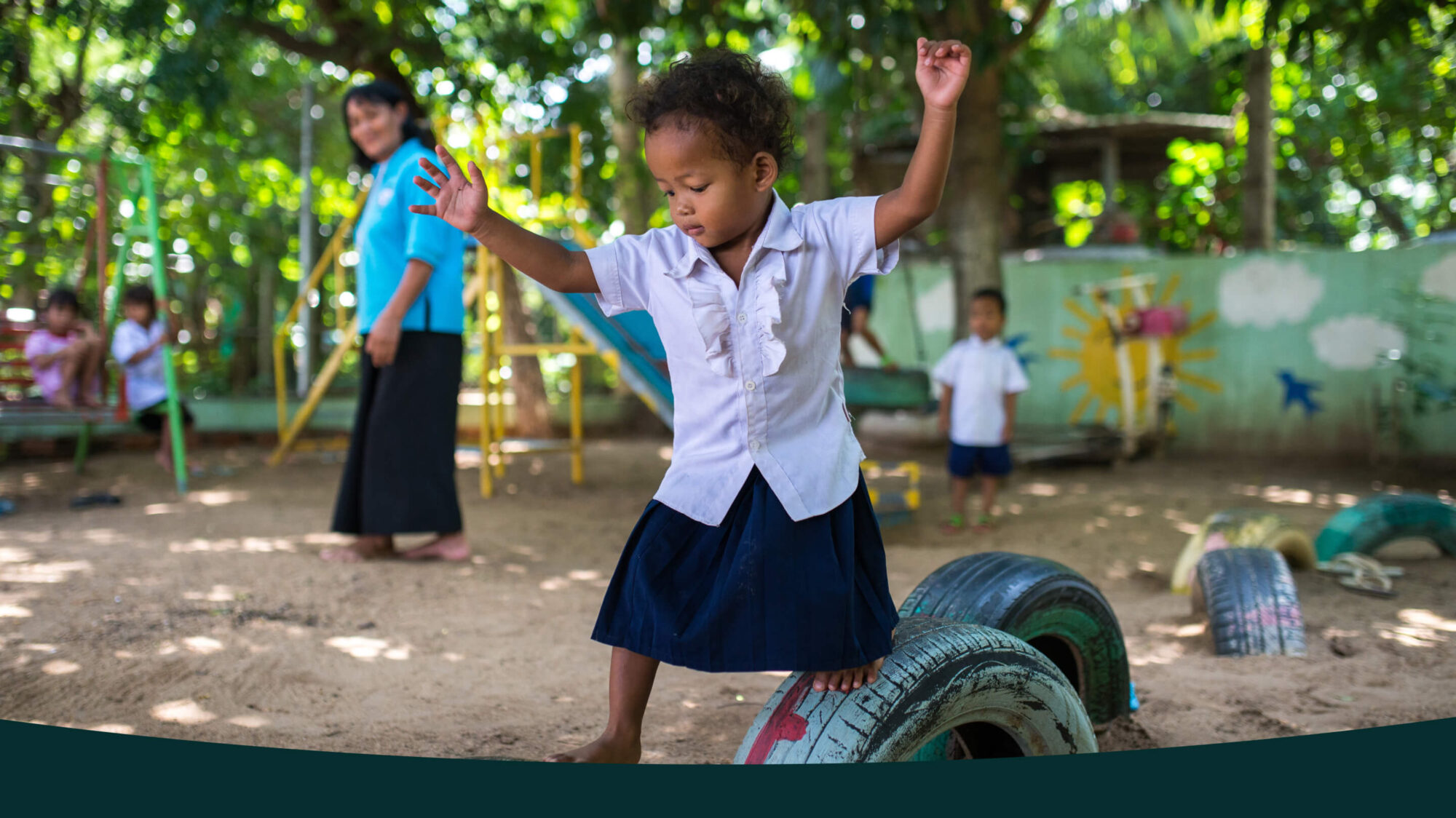 A young child in a school uniform balances on a tire in a playground while other children and an adult are in the background.