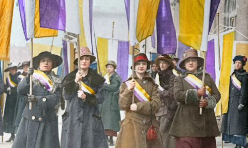 Suffragist's stand in a line, wearing long coats and hats, marching with yellow, violet, and white banners.