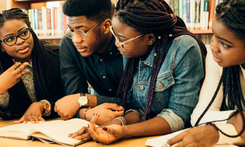 A group of black students gather around a textbook and study in the library.