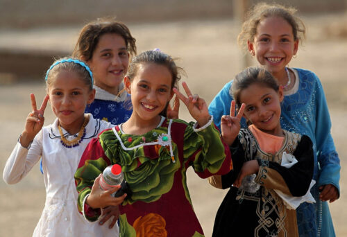 Five smiling girls standing together outdoors, holding up peace signs.