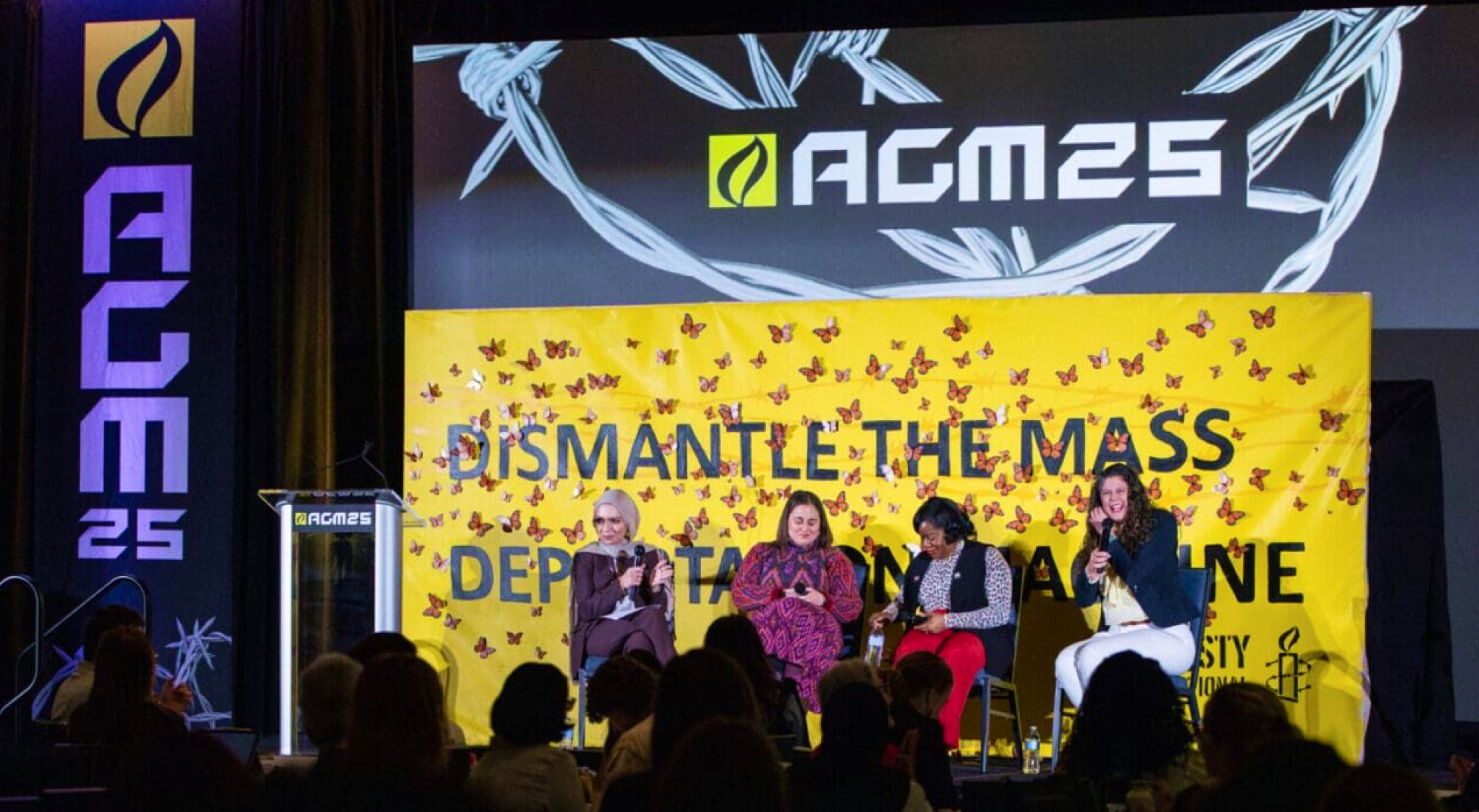 3 activists sit in front of a crowd with the AGM25 logo behind them.
