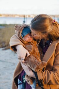 Maïca on the beach holding and kissing baby dressed in a teddy bear suit.