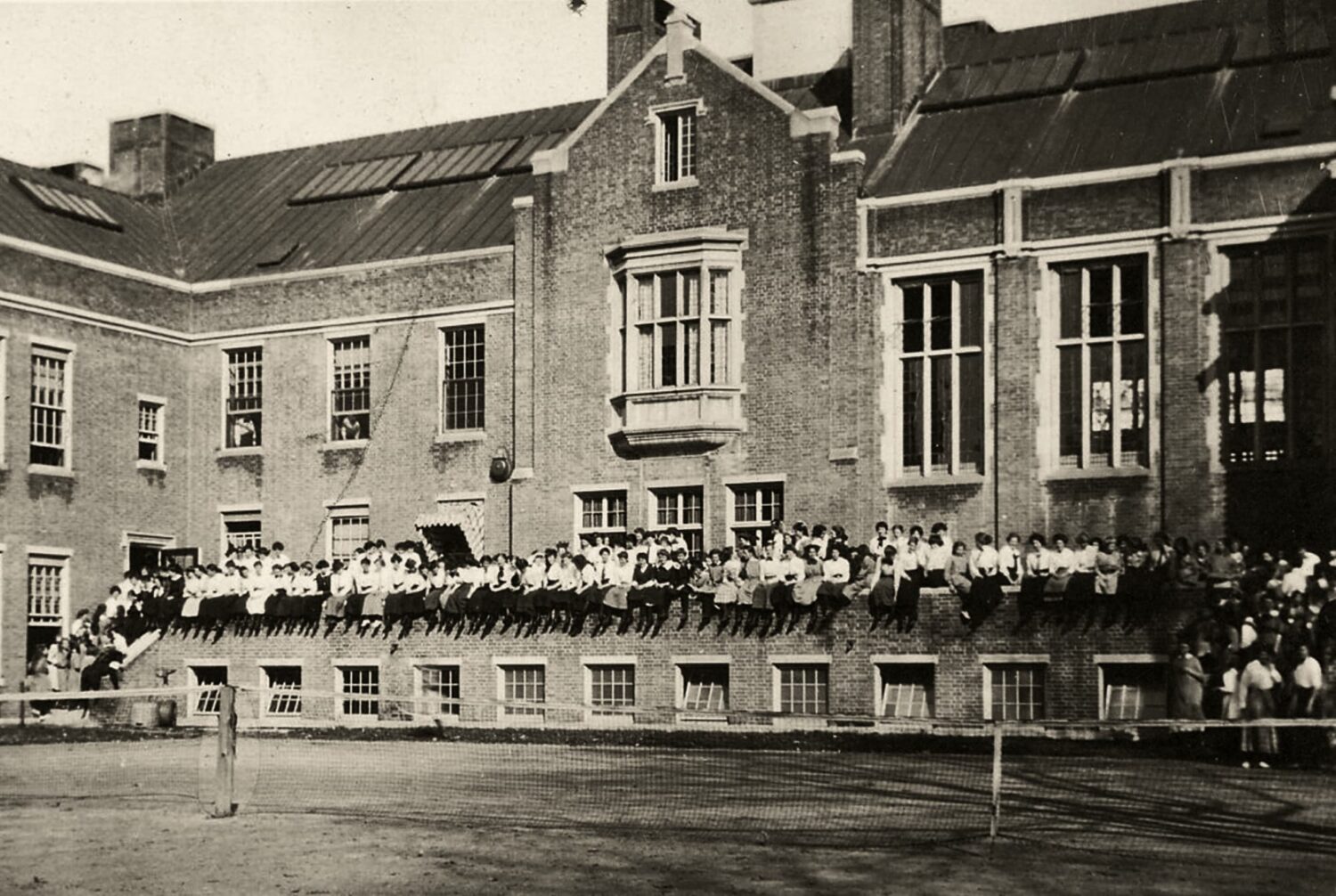 Historic sepia photo of the front of Winsor School with about 100 girls sitting on a ledge in front. Likely before the 1920s, possibly the late 1800s.