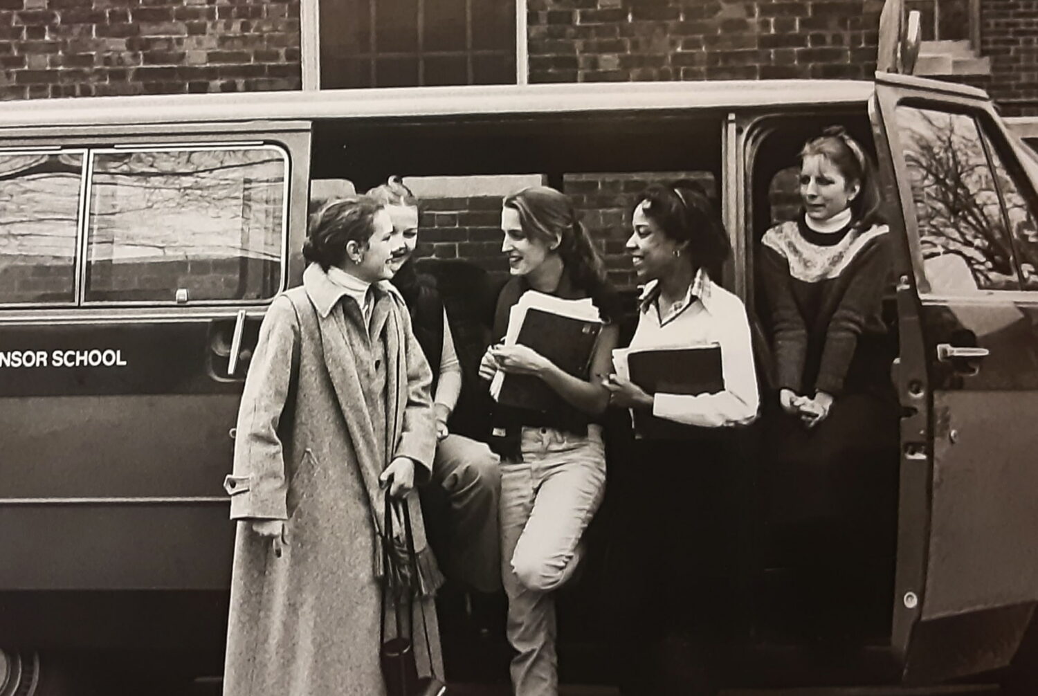 Historic sepia photo of diverse Winsor School girls standing in front of a van chatting, laughing, and holding books, likely the 1960s or 70s