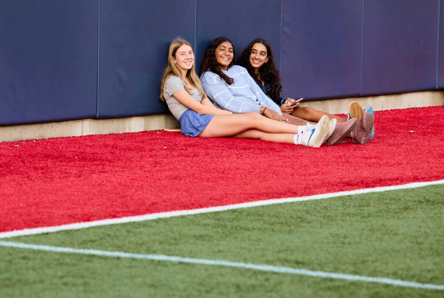 Recent image of three Winsor girls sitting in a row against a wall in on the soccer field